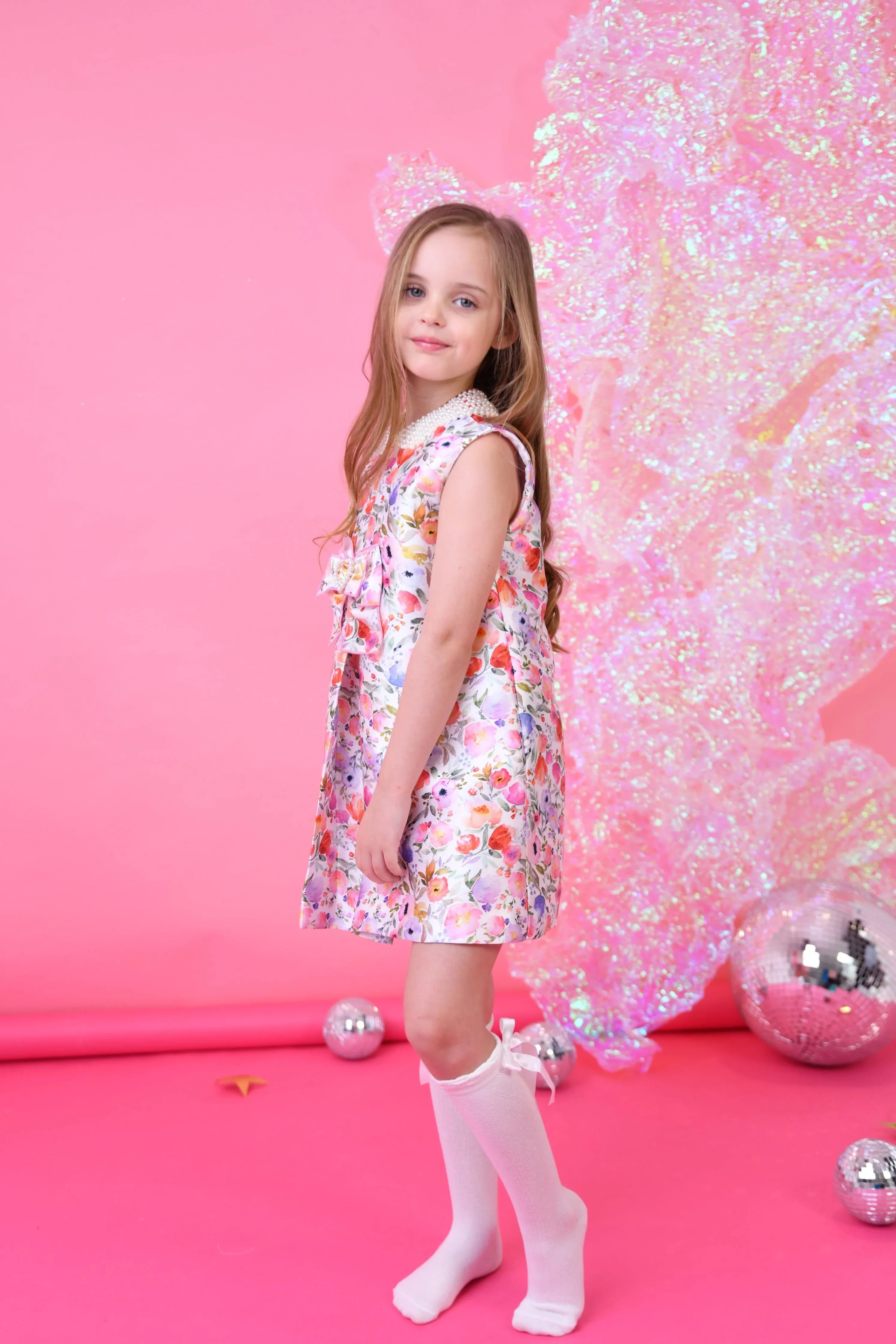 Young girl in sleeveless floral dress and white knee-high socks posing against pink backdrop with shimmering decor