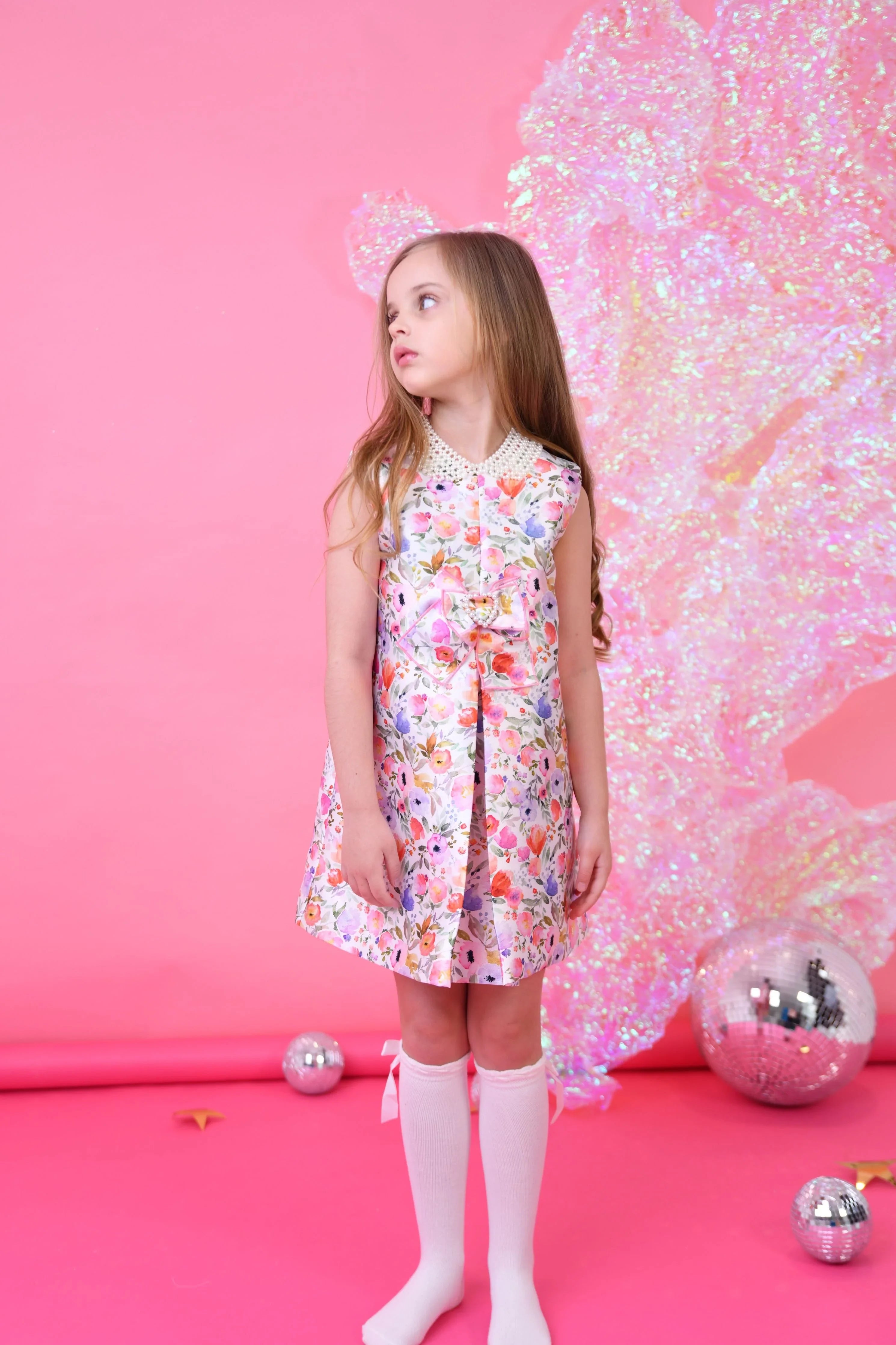 Young girl in floral dress with pearl collar and white knee-high socks against pink backdrop