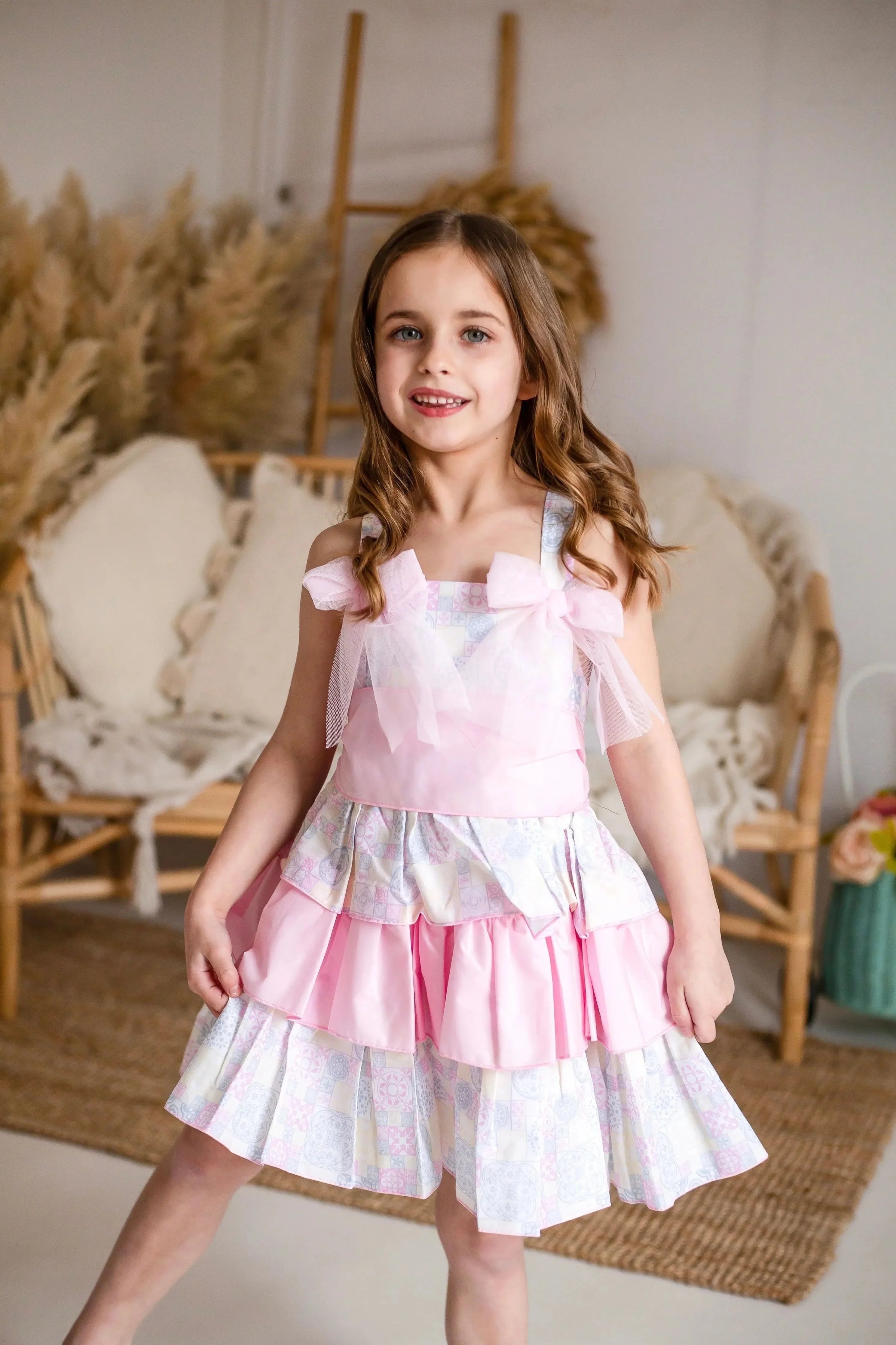 Young girl wearing a pink tiered dress with bows in a cozy room with rattan furniture and pampas grass