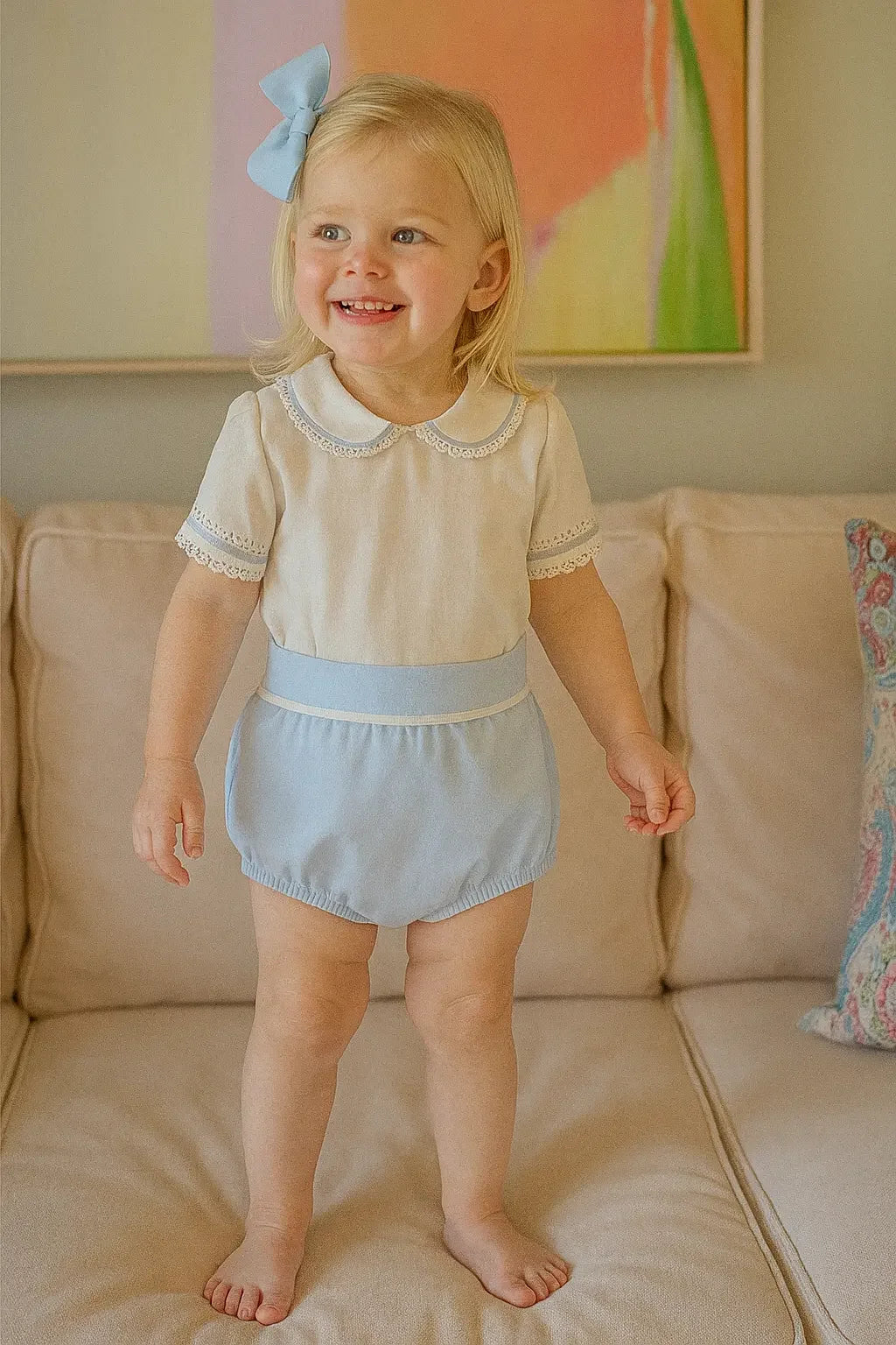 Smiling toddler girl wearing pastel blue romper with lace trim and matching hair bow on beige sofa