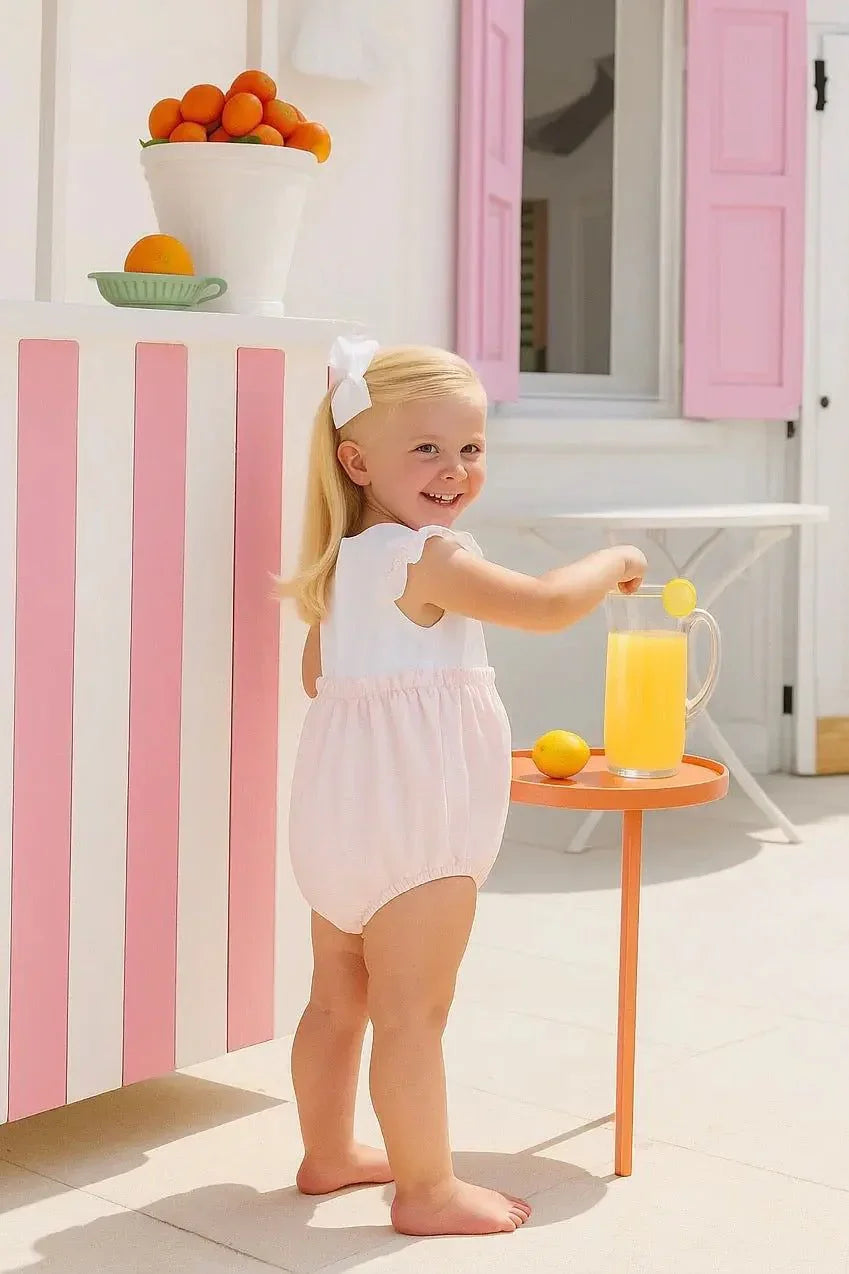Smiling toddler girl in pink ruffle romper with white bow, holding pitcher of lemonade outdoors