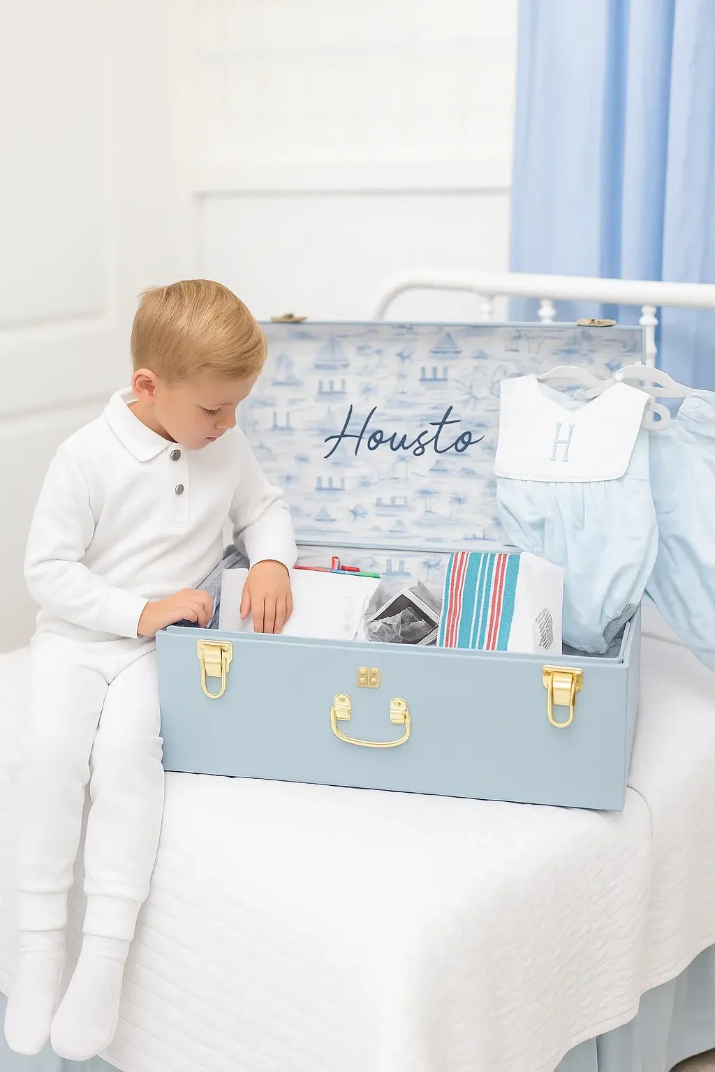 Young boy in white pajamas sitting on bed next to light blue personalized toy chest with clothes