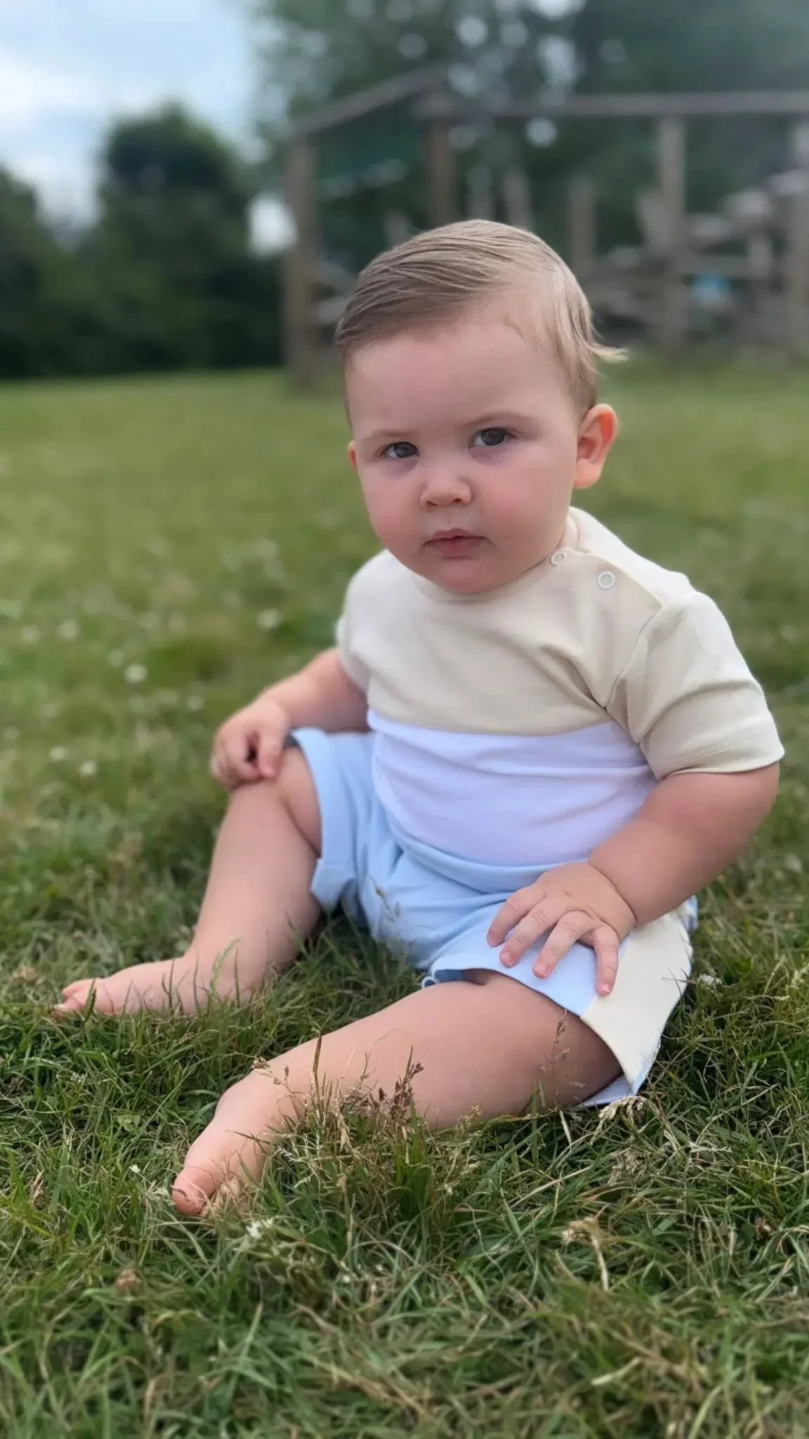 Baby boy sitting on grass outdoors wearing cream and white top with light blue shorts