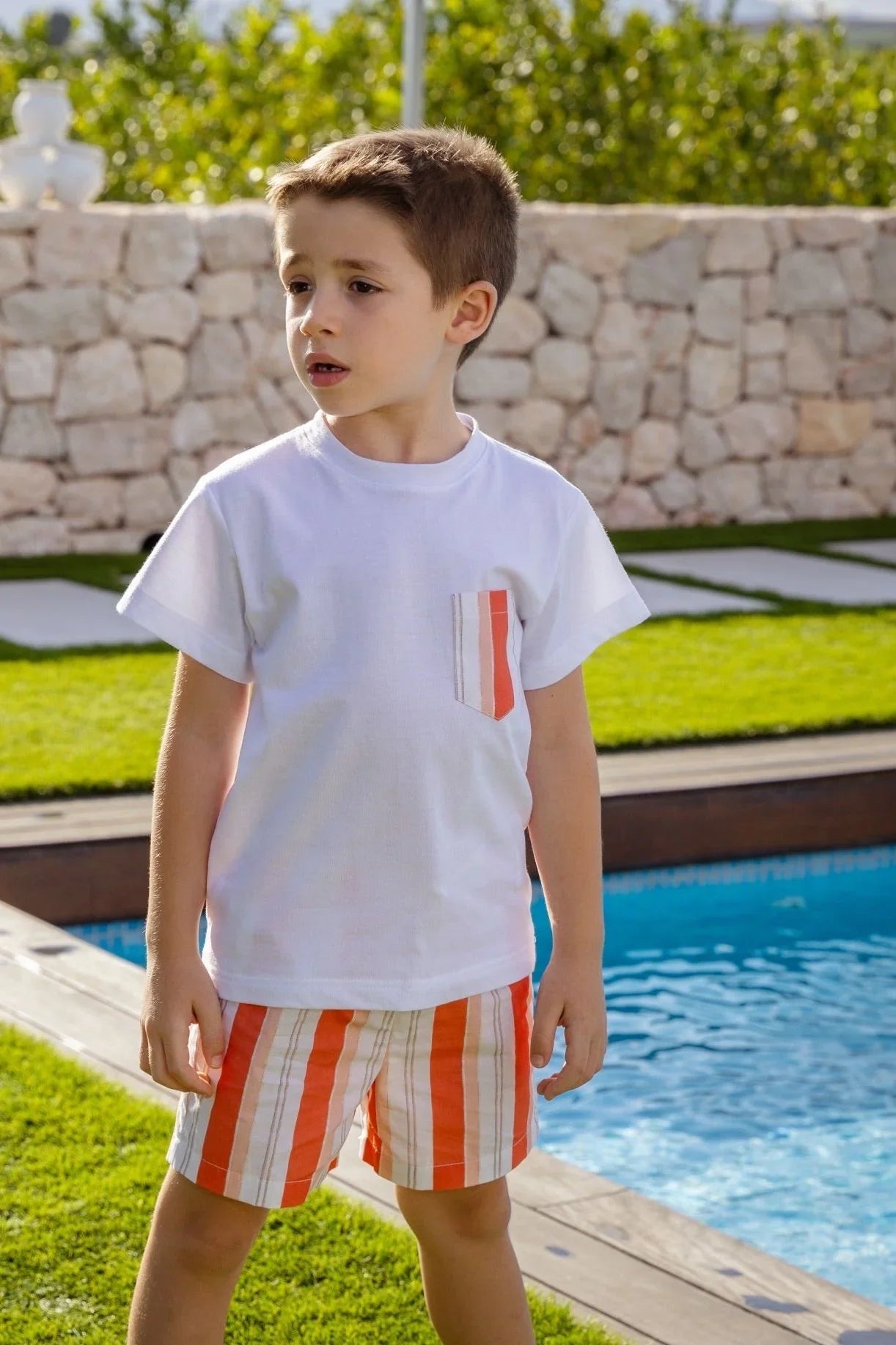 Young boy wearing white t-shirt with striped pocket and orange striped swim shorts by pool