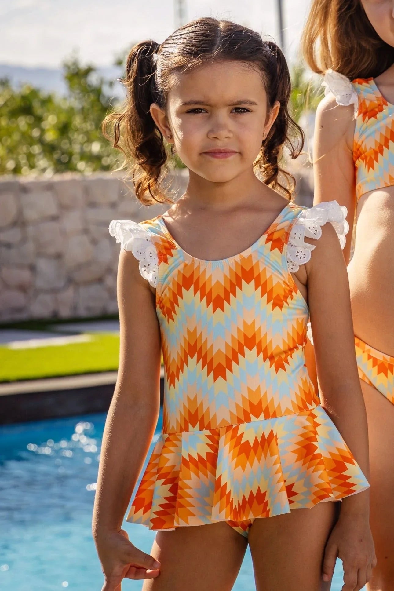 Young girl wearing orange and blue patterned swimsuit with ruffle sleeves by pool on sunny day