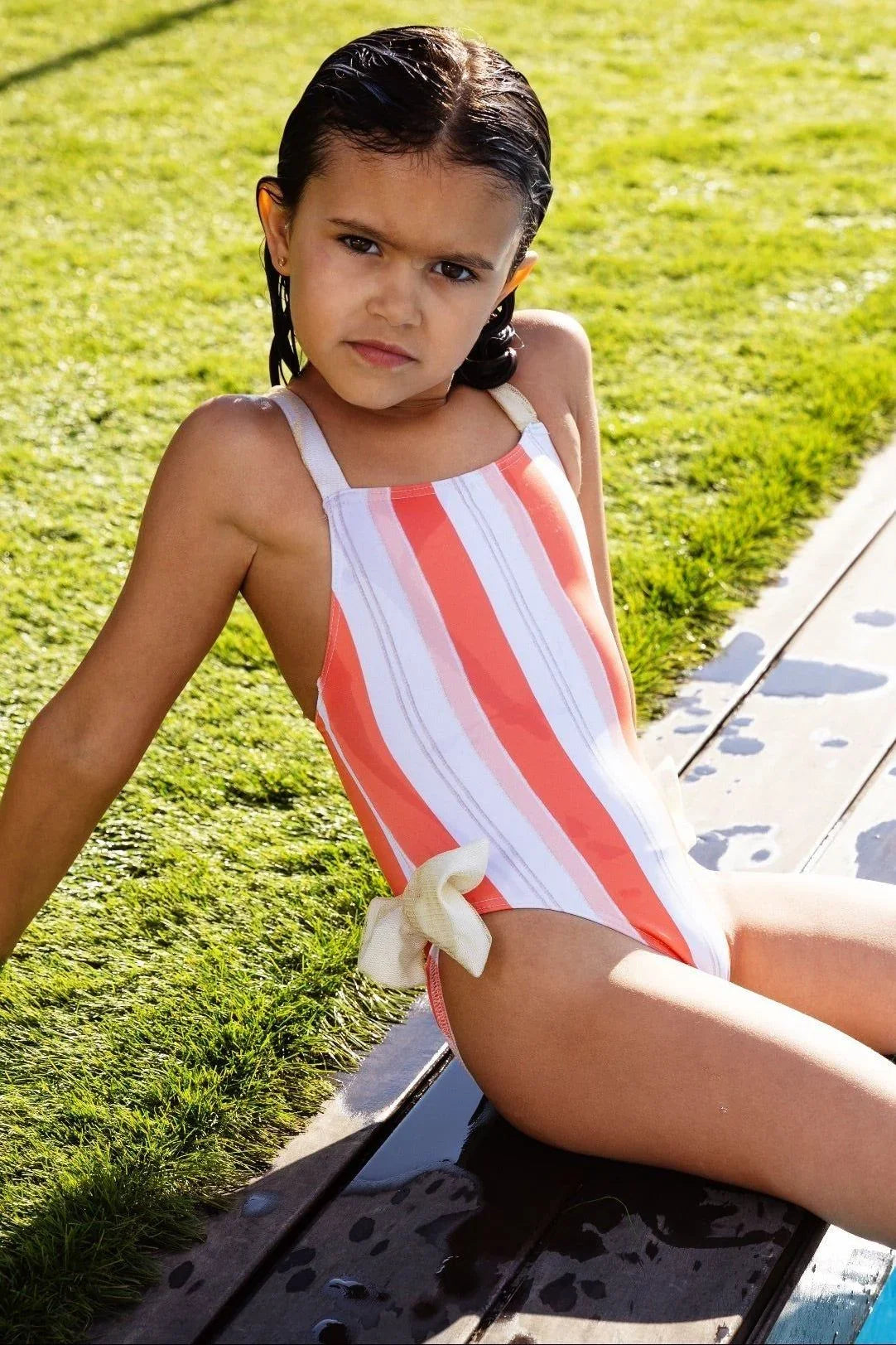 Young girl in orange and white striped swimsuit with bow detail sitting by pool on sunny grass