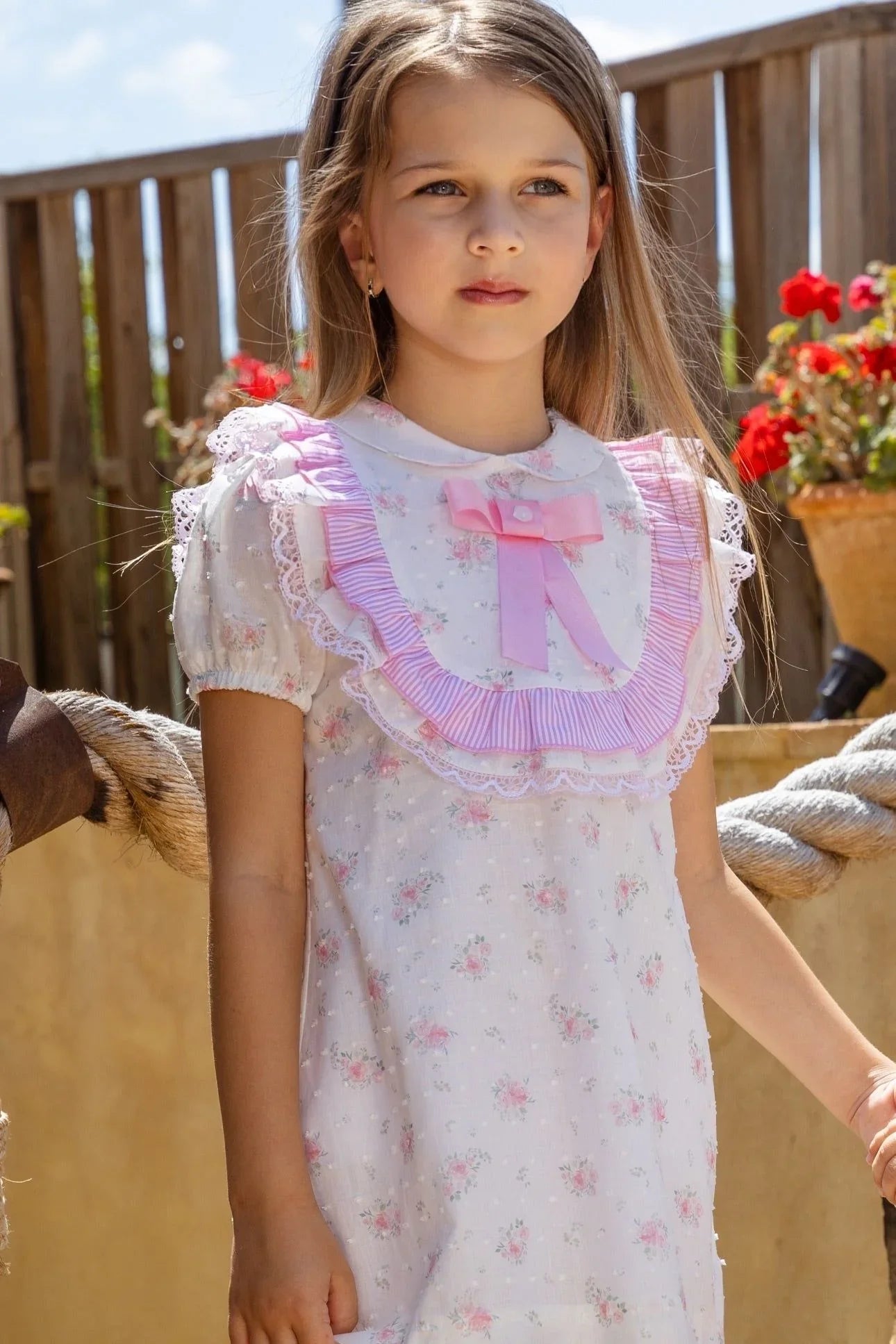 Young girl wearing a pink floral dress with ruffled collar and bow, outdoor sunny setting