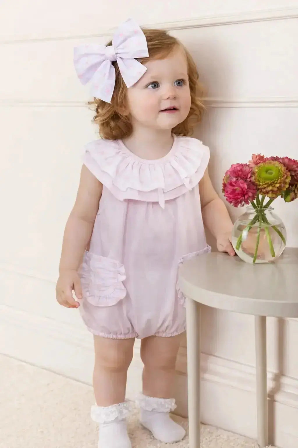 Young child in a pink outfit with a large bow, standing next to a small table with a vase of flowers.