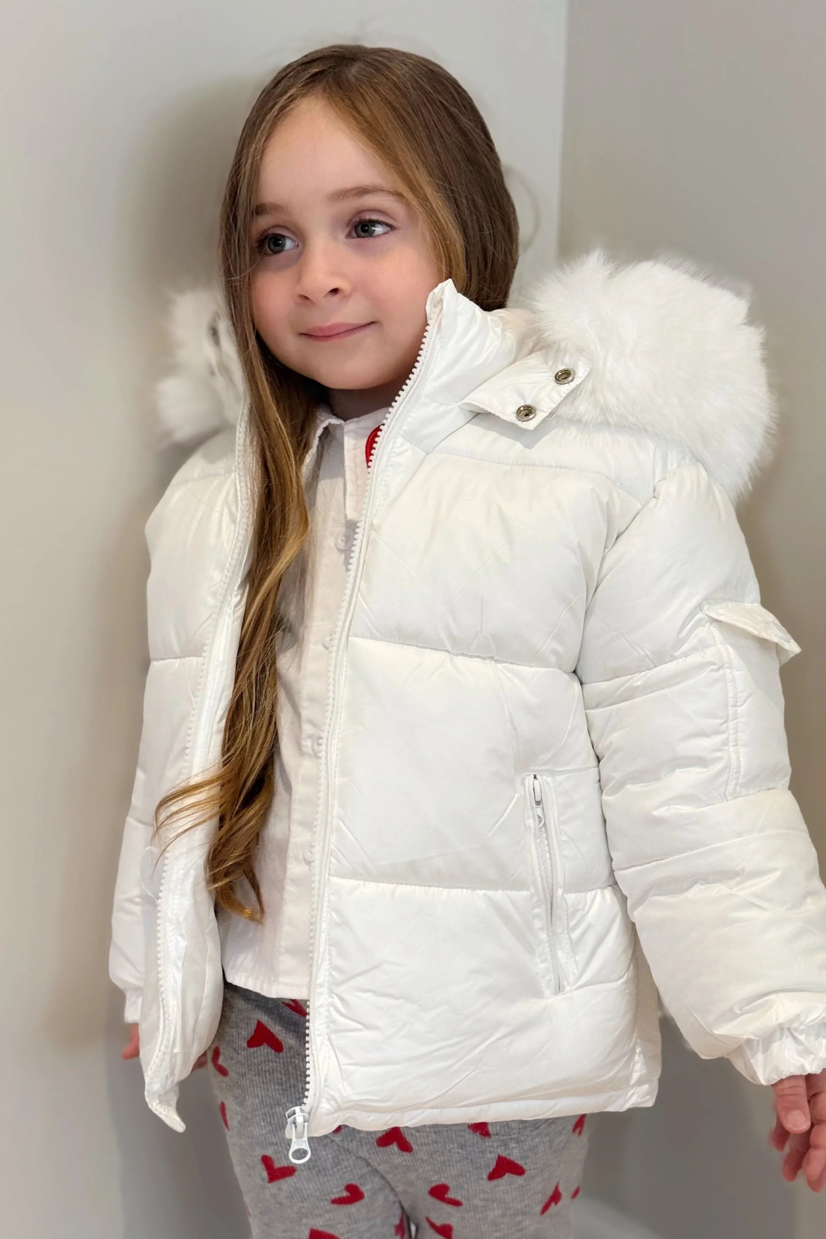 Young girl in white puffer jacket with fur hood, gray pants with red hearts, standing indoors