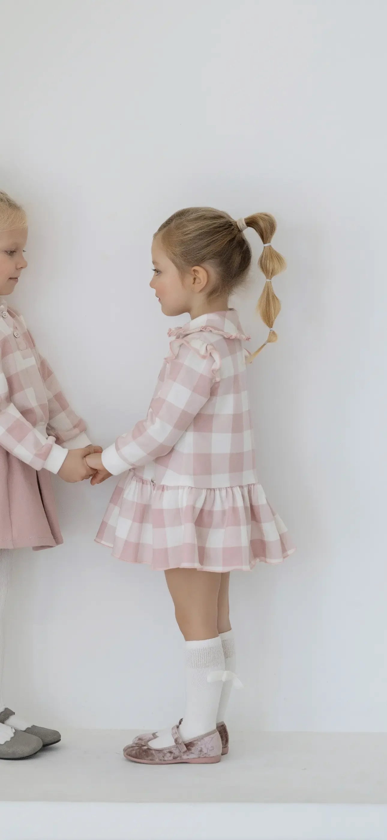 Two young girls holding hands wearing pink and white checkered dresses and white socks with bows