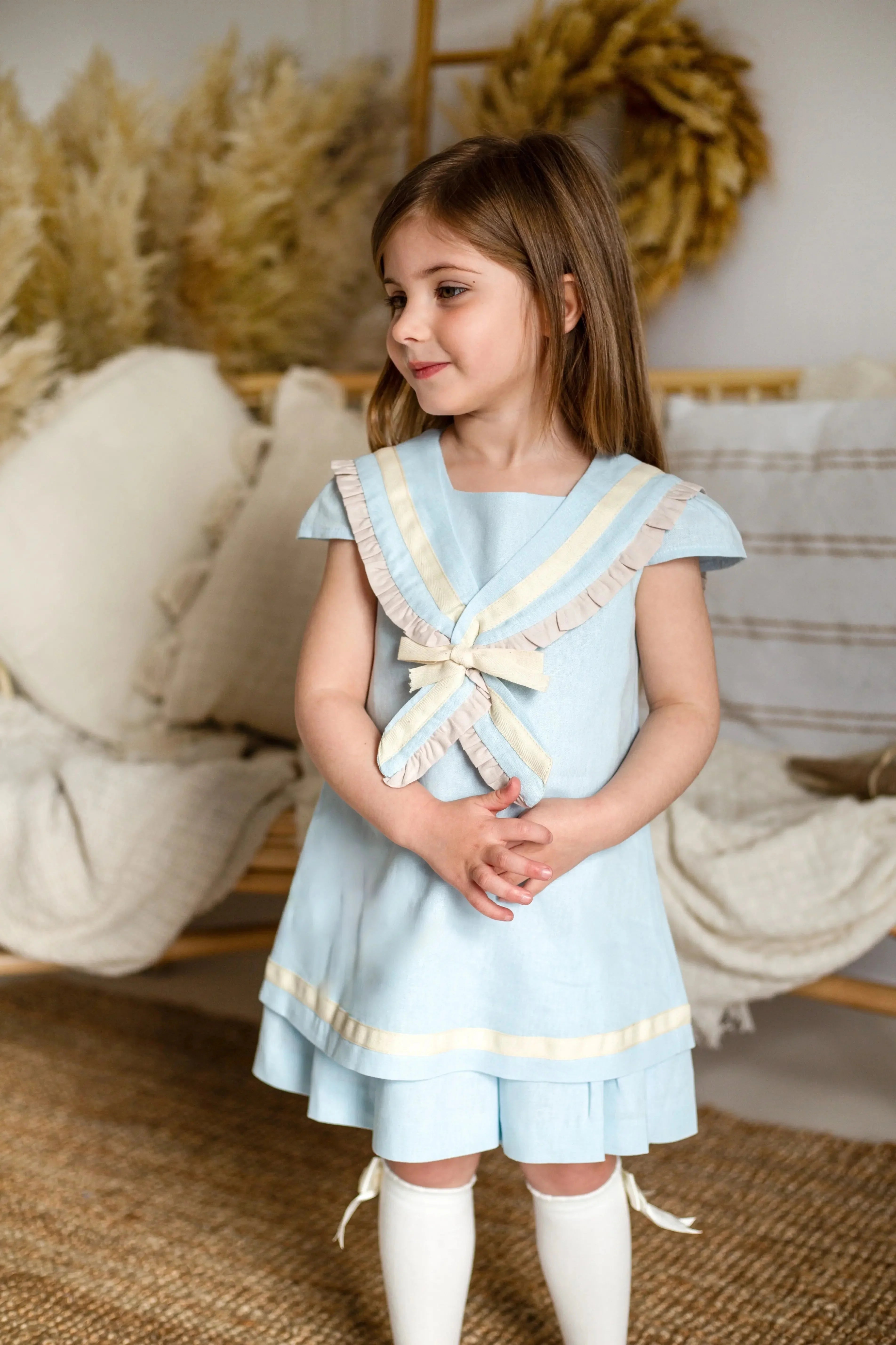 Little girl wearing a light blue sailor linen dress with cream trim, standing indoors by cozy cushions