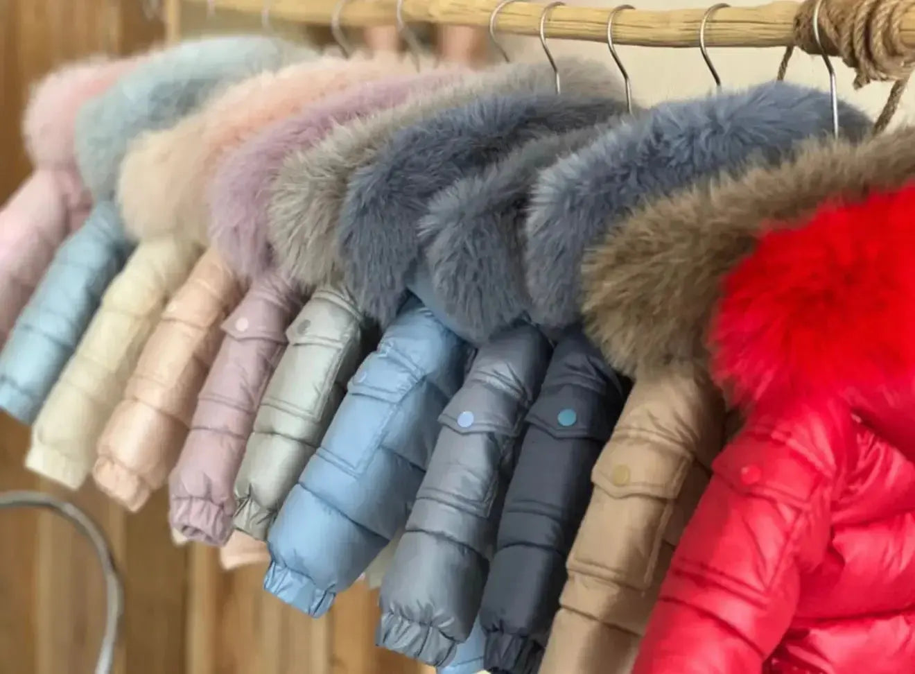 Colorful winter coats with fur hoods hanging on a wooden rack, displaying pastel and bright shades