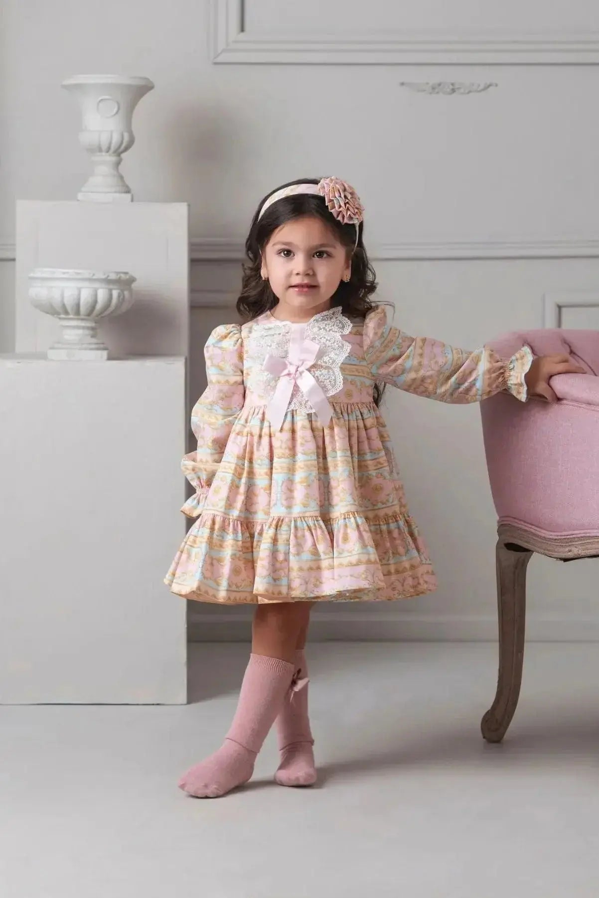 Young girl in pastel floral dress with lace and bow, pink headband, and knee-high socks leaning on pink chair