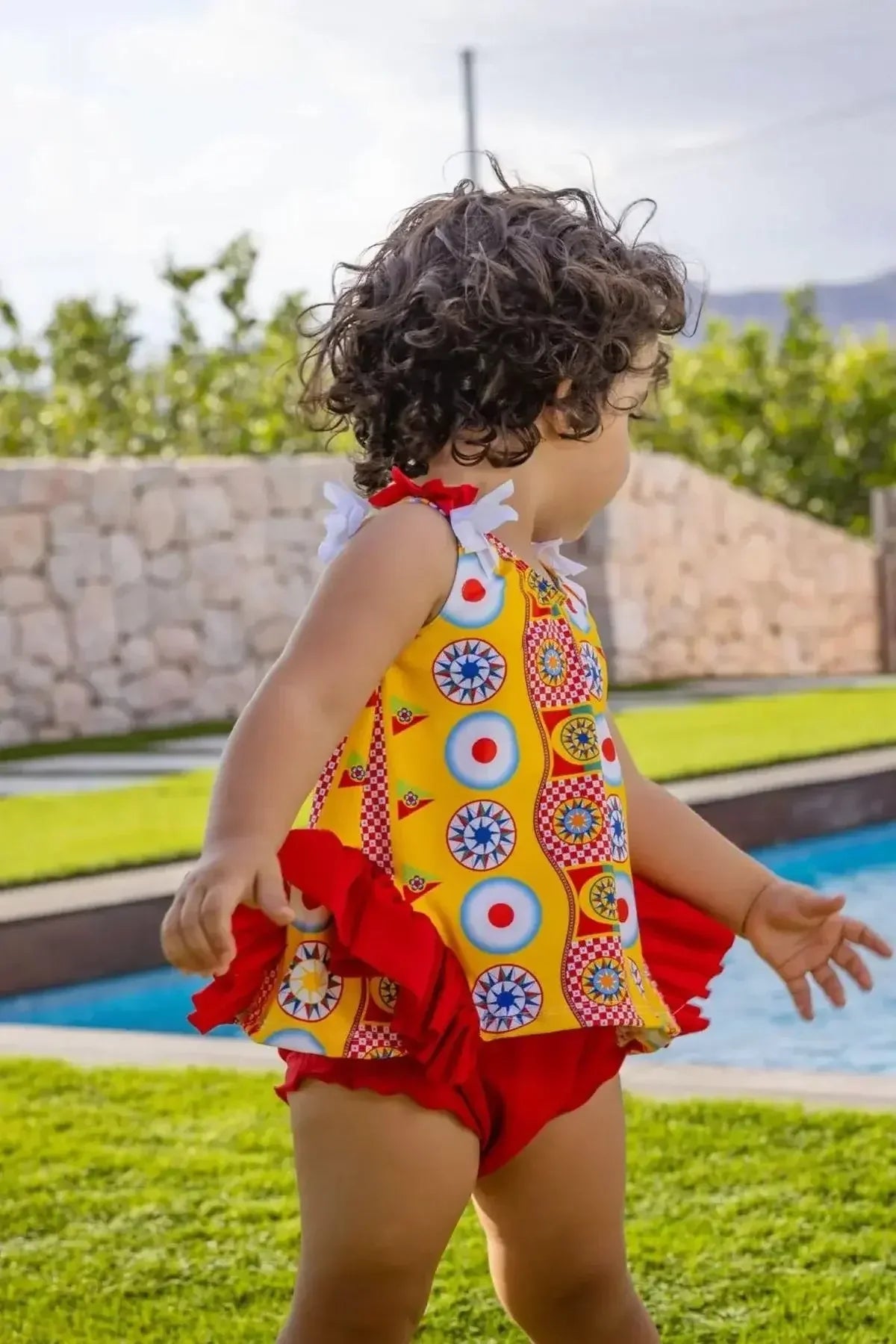 Toddler wearing a colorful patterned yellow and red swimsuit standing near a pool in a backyard