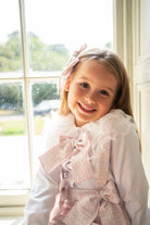 Smiling young girl wearing a white blouse with ruffled collar and pink bow accessories by a window