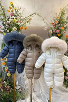 Three baby winter snowsuits in blue, beige, and white with fur-trimmed hoods displayed on stands