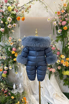 Blue child's puffer snowsuit with fur trim on collar and cuffs displayed on mannequin amidst floral decor
