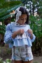 Girl wearing blue ruffled top with carousel print shorts and matching bow headband in garden