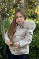 Young girl wearing a beige puffer jacket with fur hood, holding a green leaf outdoors
