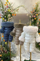Three puffer winter jumpsuits with fur collars and cuffs on gold mannequins, surrounded by floral arrangements