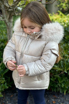 Child in a beige puffer jacket with fur hood standing outdoors among green plants