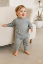 Smiling toddler standing barefoot in light gray knit sweater and pants indoors near beige armchair
