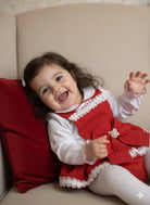 Happy toddler girl in red dress with white lace sitting on beige couch with red pillow