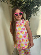 Girl wearing yellow lemon and pink bow print outfit and pink heart-shaped sunglasses standing by potted plant