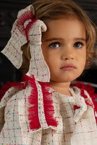Close-up of a toddler with brown eyes wearing a textured white outfit with red ruffle details and matching large bow