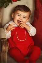 Smiling baby with blue eyes wearing red romper and white shirt sitting on beige chair with red cushion