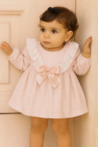 Toddler girl in pink dress with ruffles and bow, standing indoors by light beige wall