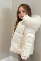 Young girl wearing a white puffer winter jacket with fur hood, standing indoors against neutral wall