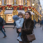 Woman in dark fur coat holding child in light blue puffer jacket on festive city street with holiday lights