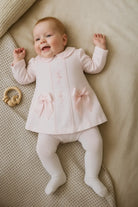 Smiling baby in pink dress with bows and white tights lying on beige textured blanket with wooden rattle