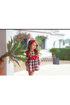 Young girl in red and white plaid dress with red bow headband outdoors near large potted plant