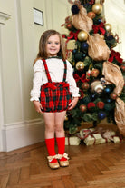 Little girl in red and green Christmas tartan romper with red socks and gold shoes standing by decorated Christmas tree