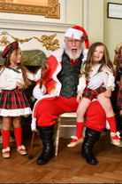 Santa Claus in red suit with two girls wearing Christmas tartan outfits and red socks in festive indoor setting