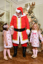 Santa Claus in red suit holding hands with two girls in pink floral dresses and knee-high socks near Christmas tree