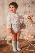 Toddler boy in ruffled white blouse and floral romper with knee socks standing on wooden floor