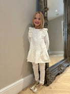 Smiling girl in cream pearl embellished tunic with ruffles and leggings, wearing gold shoes indoors