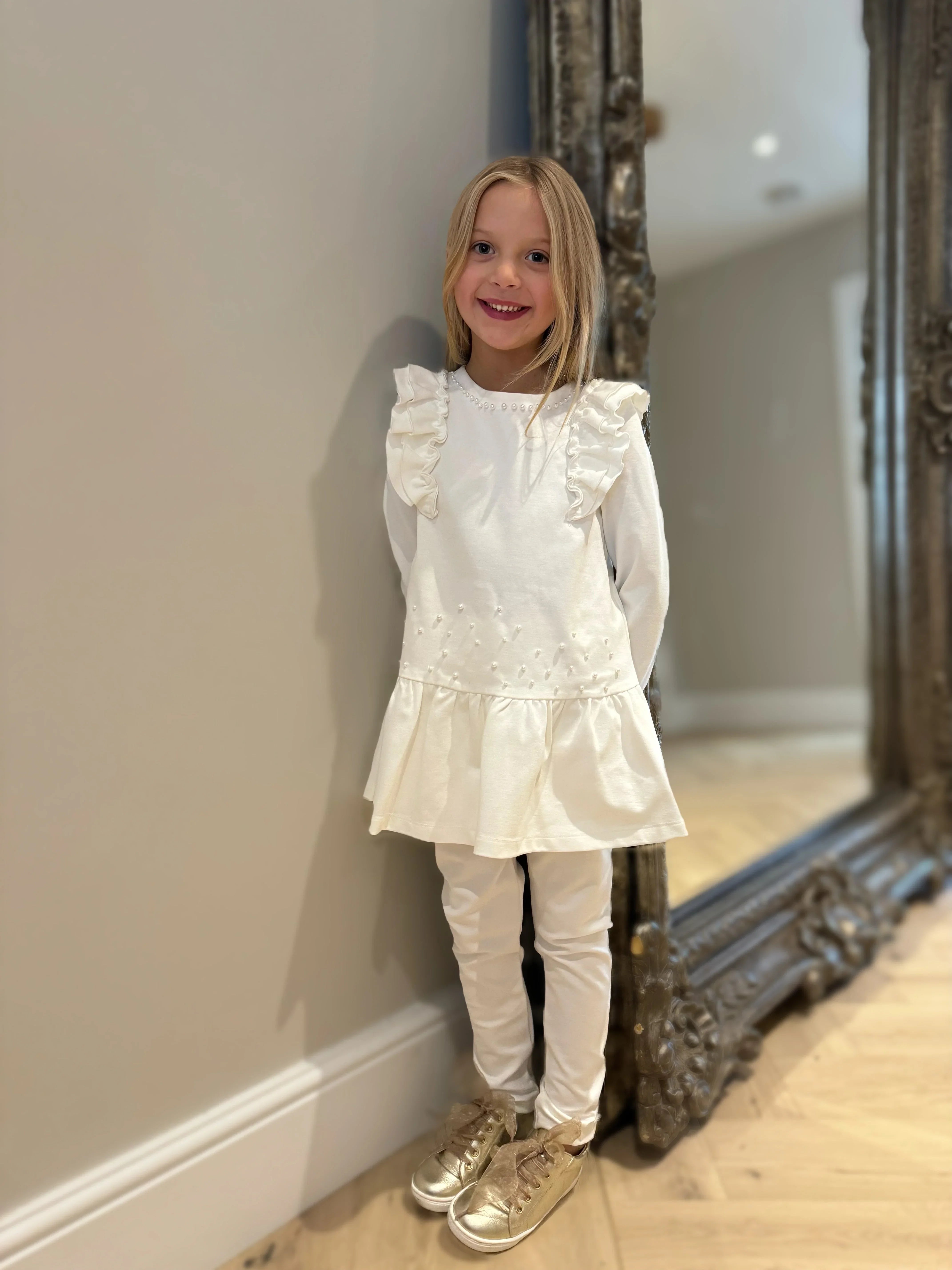 Smiling girl in cream pearl embellished tunic with ruffles and leggings, wearing gold shoes indoors
