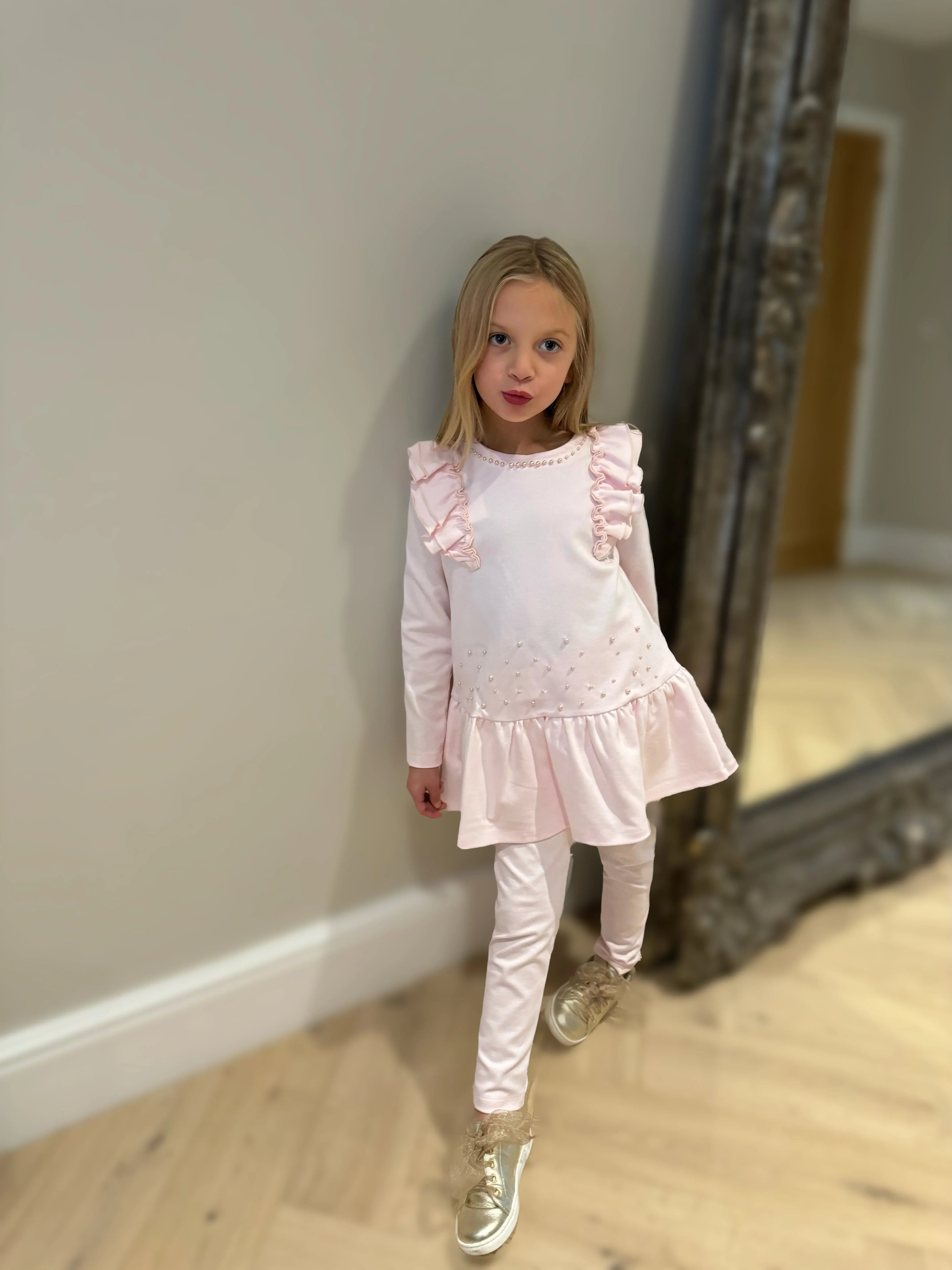 Young girl in pink ruffled long-sleeve dress and leggings posing indoors with gold sneakers