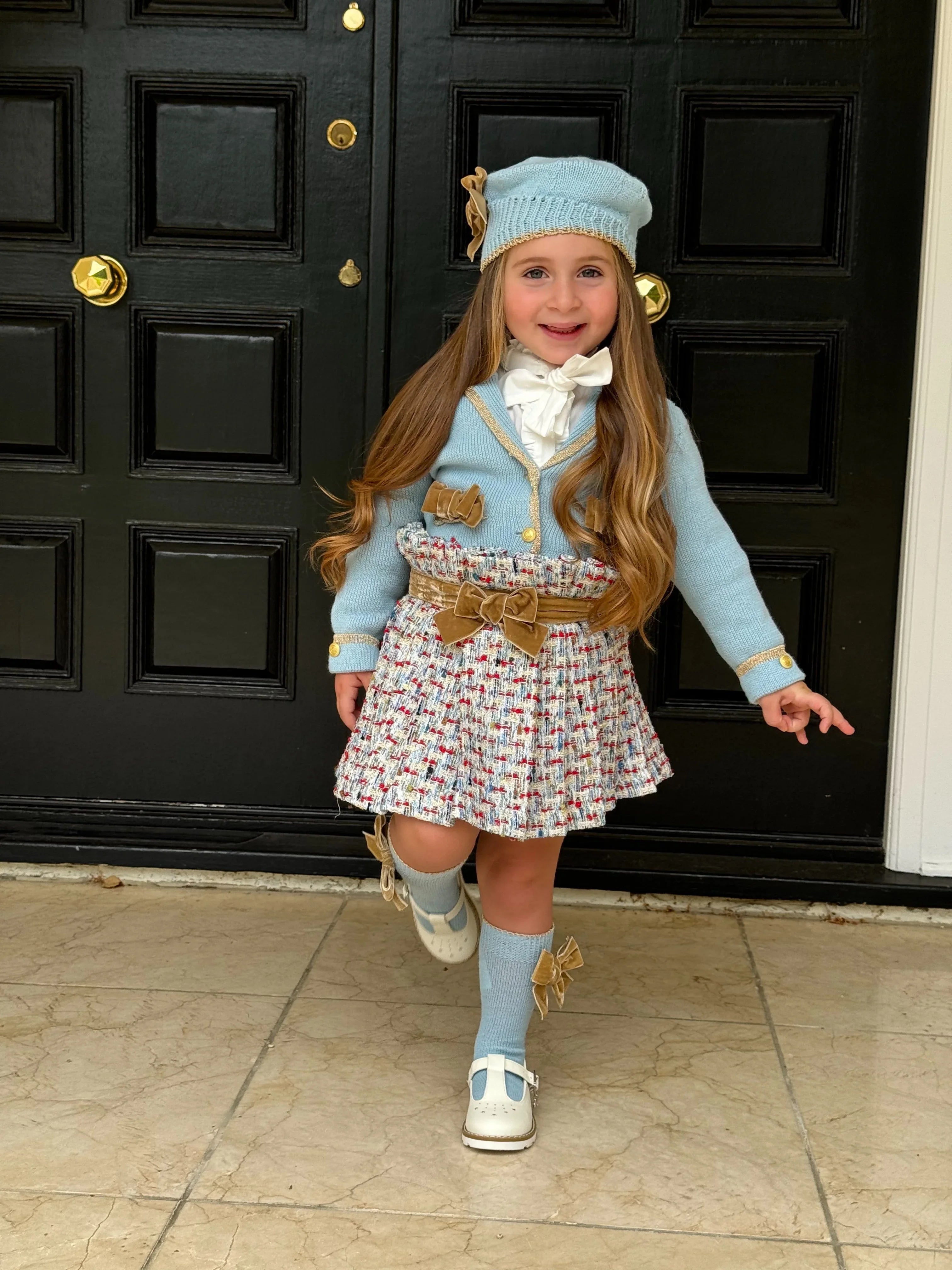 Young girl in blue knit hat, matching cardigan, plaid skirt with bows, standing on tiled floor