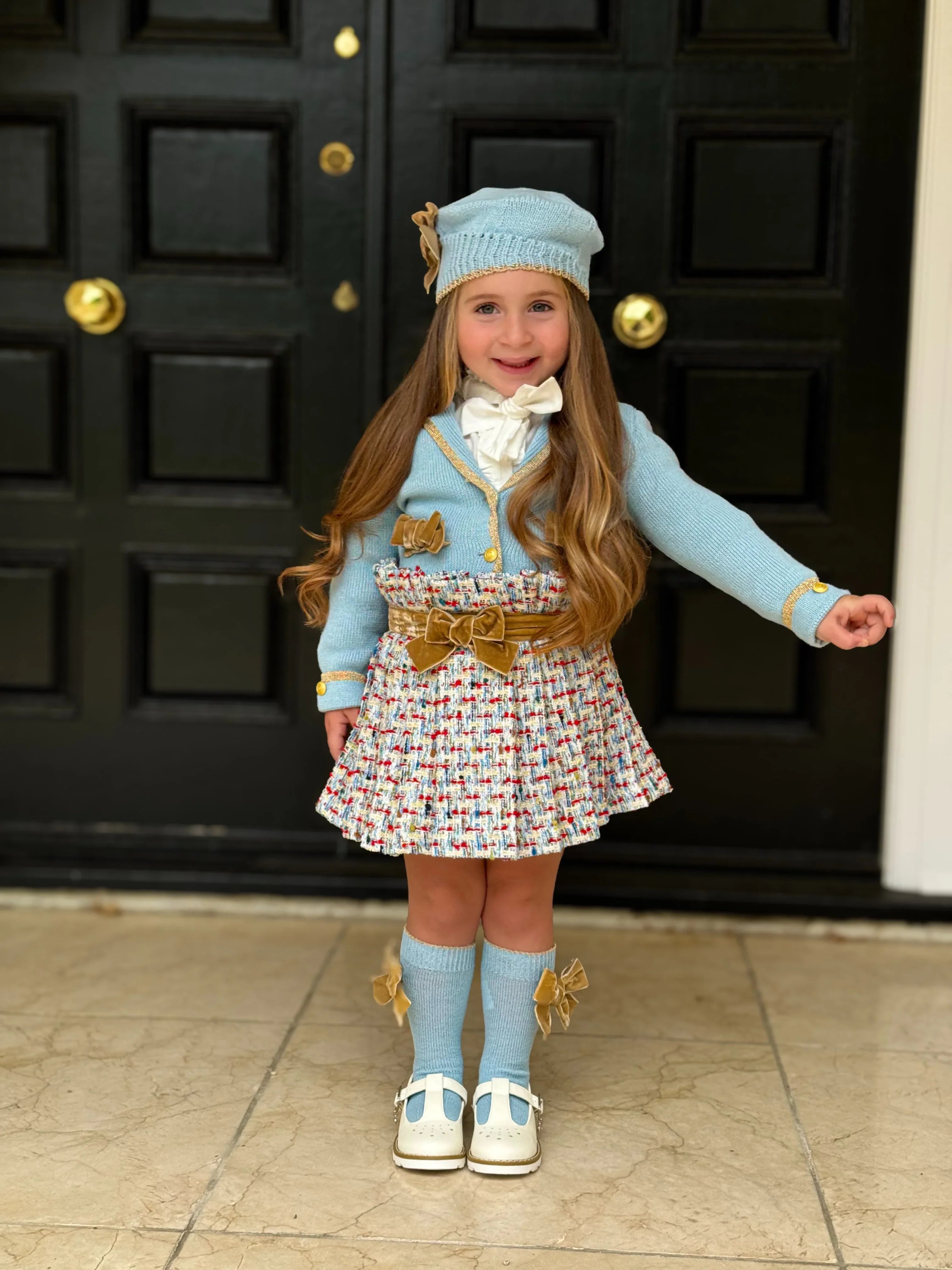 Little girl in stylish blue knit hat, sweater with gold accents, tweed skirt, knee-high socks, and white shoes