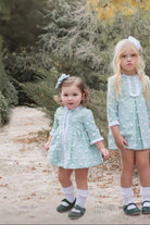 Two young girls wearing matching light green textured dresses with white frilled details and bows, standing outdoors on a path surrounded by greenery