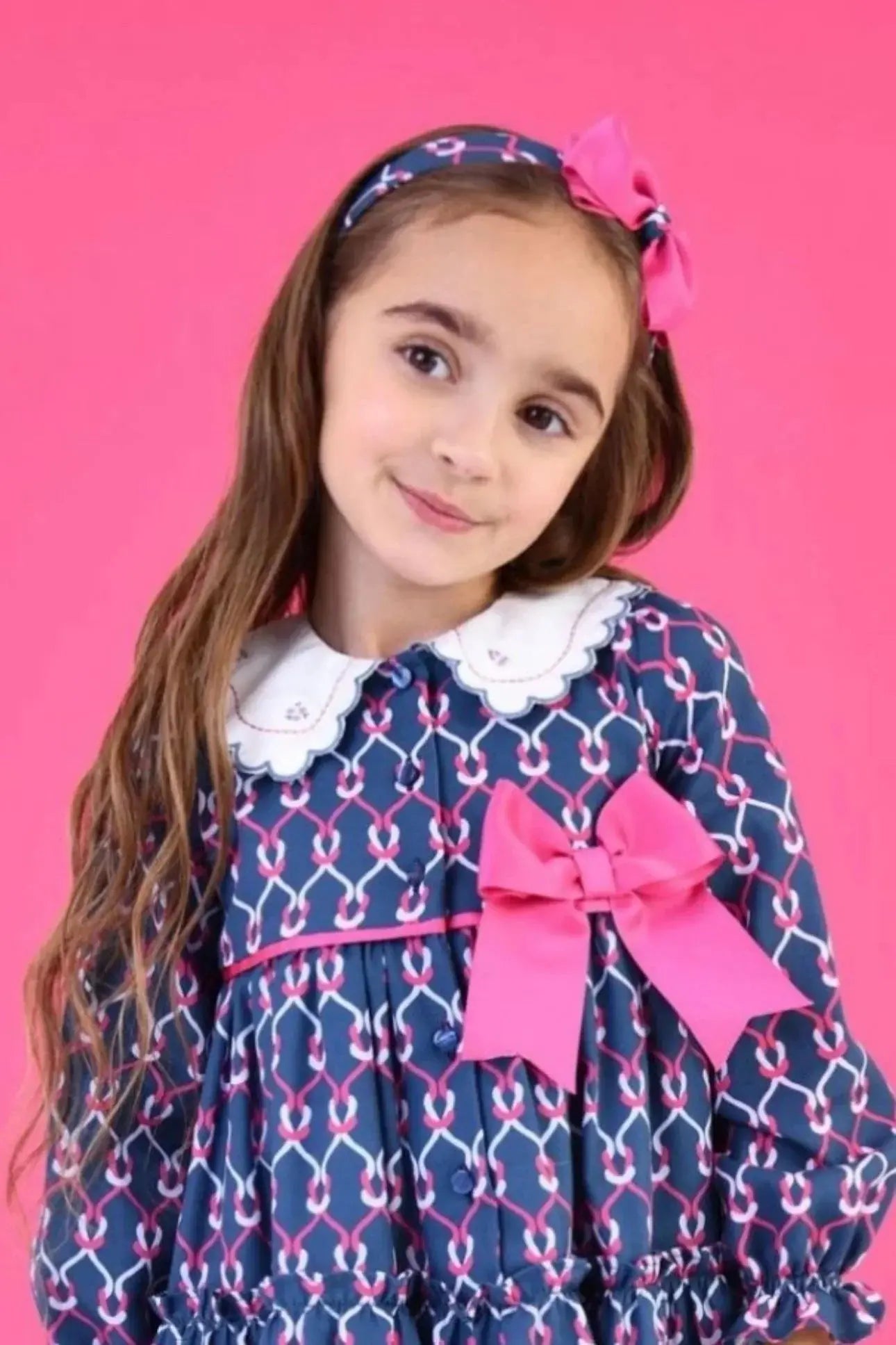 Young girl in blue patterned dress with white collar and pink bow, wearing matching headband on pink background