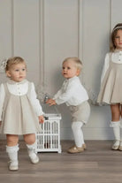 Three toddlers wearing beige pinafore dresses and white outfits, posing indoors with decorative birdcage
