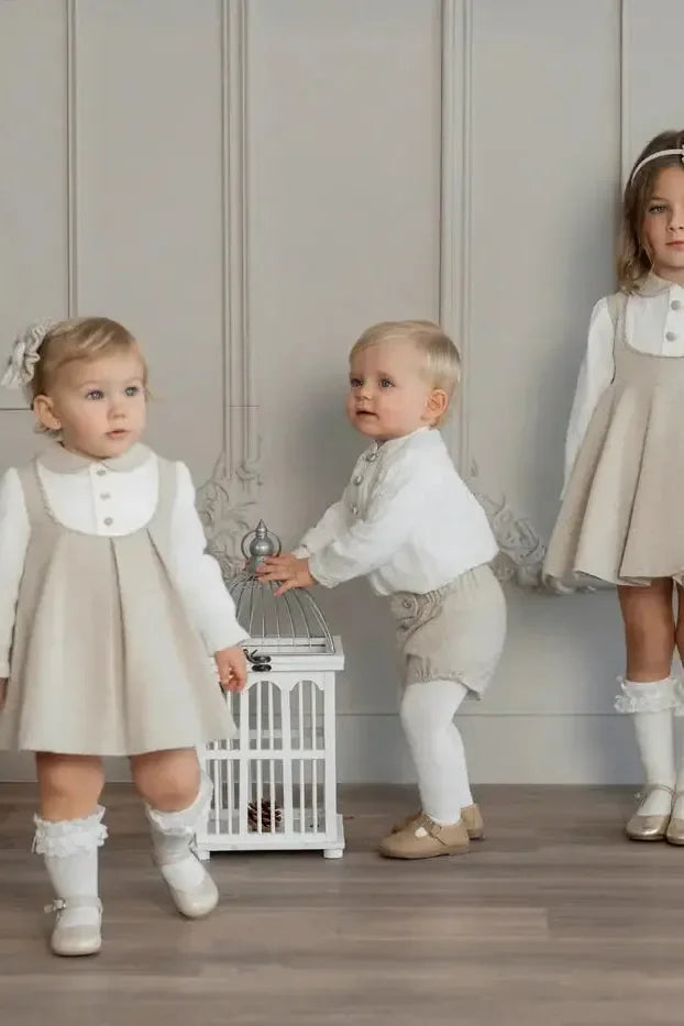 Three toddlers wearing beige pinafore dresses and white outfits, posing indoors with decorative birdcage