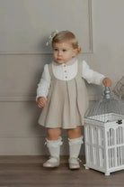 Toddler girl in beige pinafore dress with white blouse and bow headband standing by decorative white birdcage