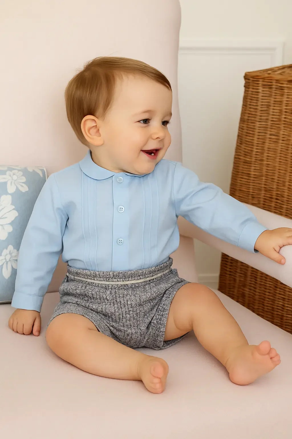Smiling baby boy sitting on a pink armchair wearing a light blue shirt and gray tweed shorts
